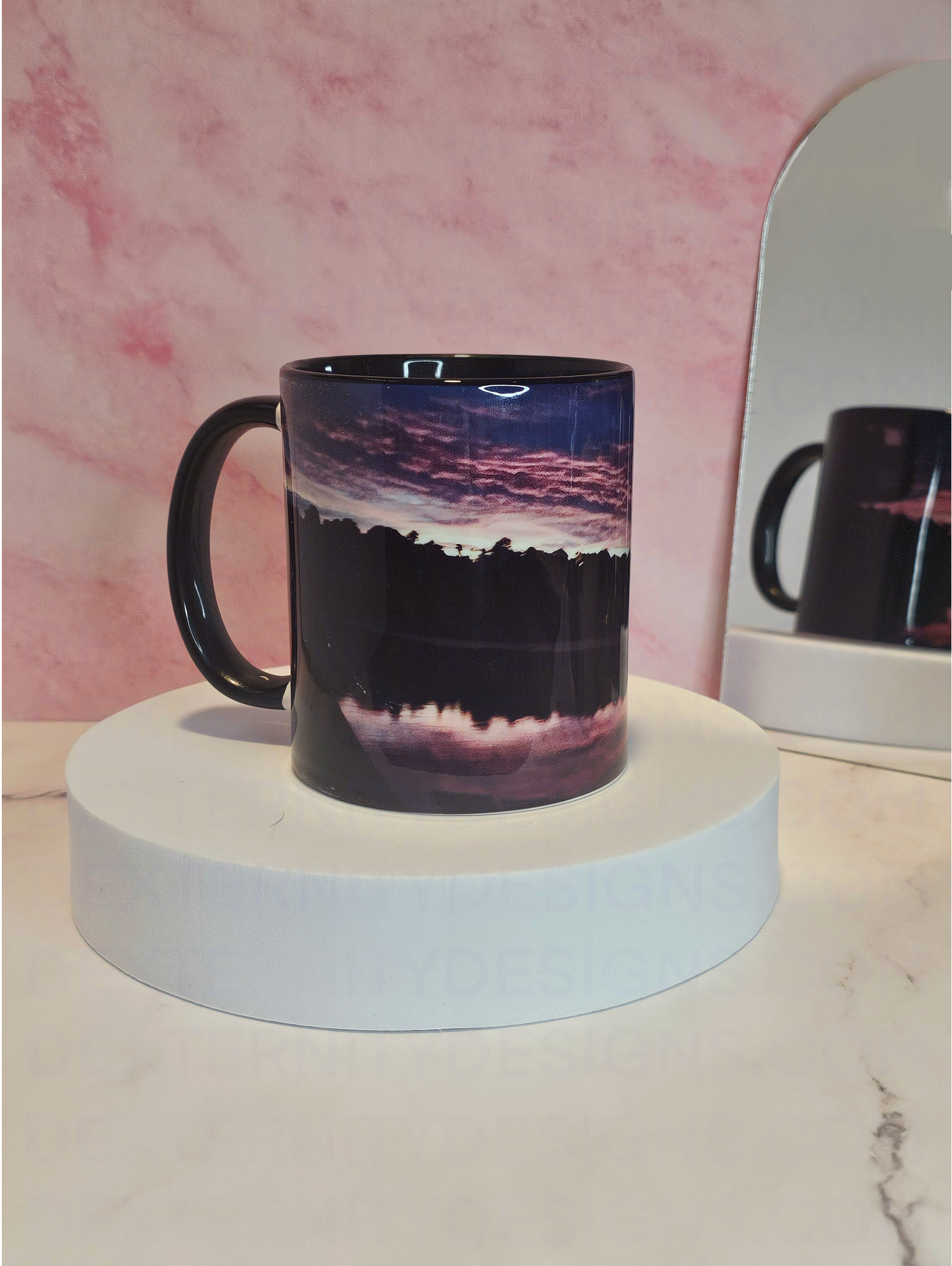 Mug with a scenic sunrise design on a white platform against a pink marble background with a mirror reflecting it's back behind it.