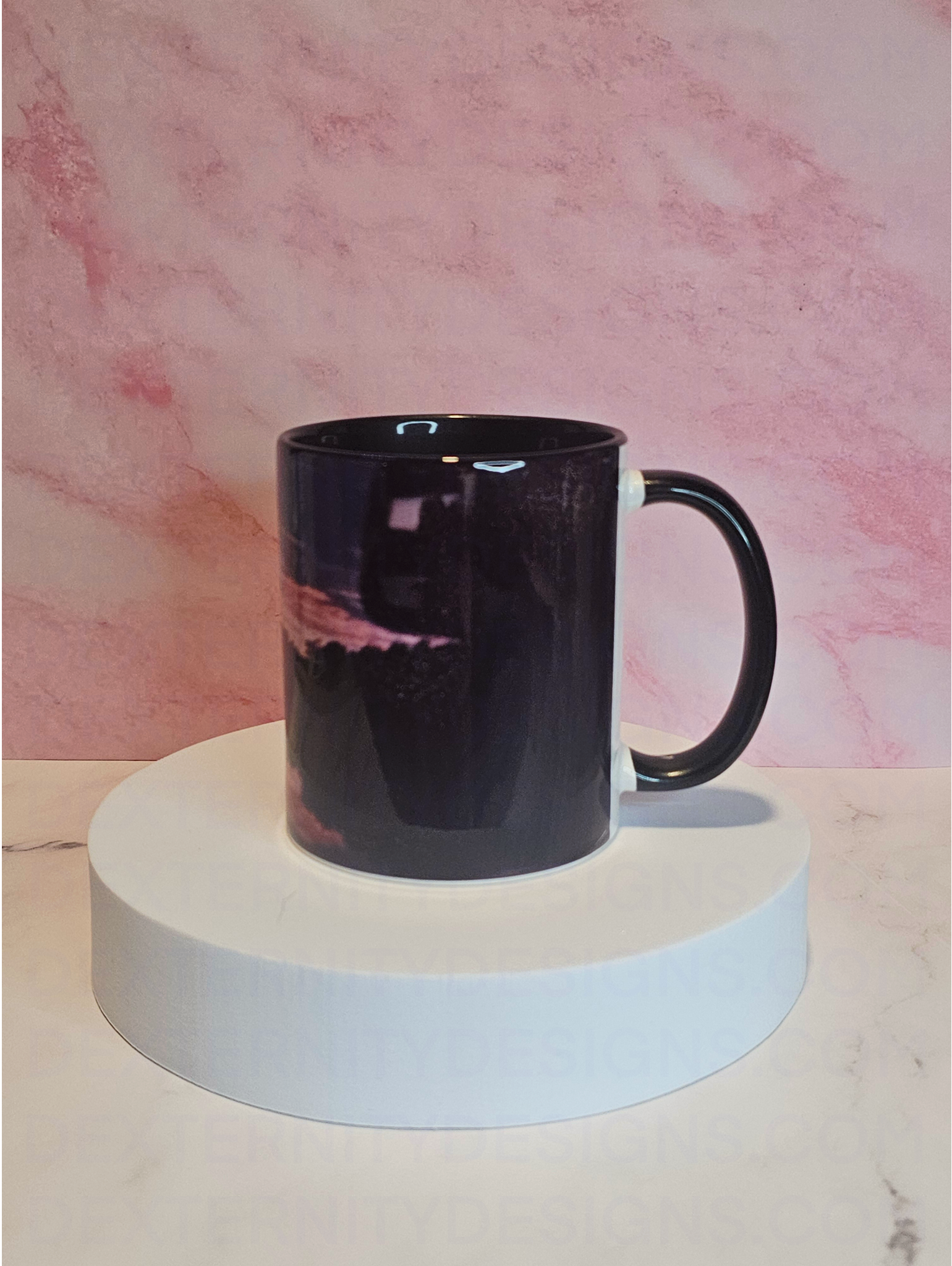 Side view of a mug with a scenic sunrise design on a white platform against a pink marble background