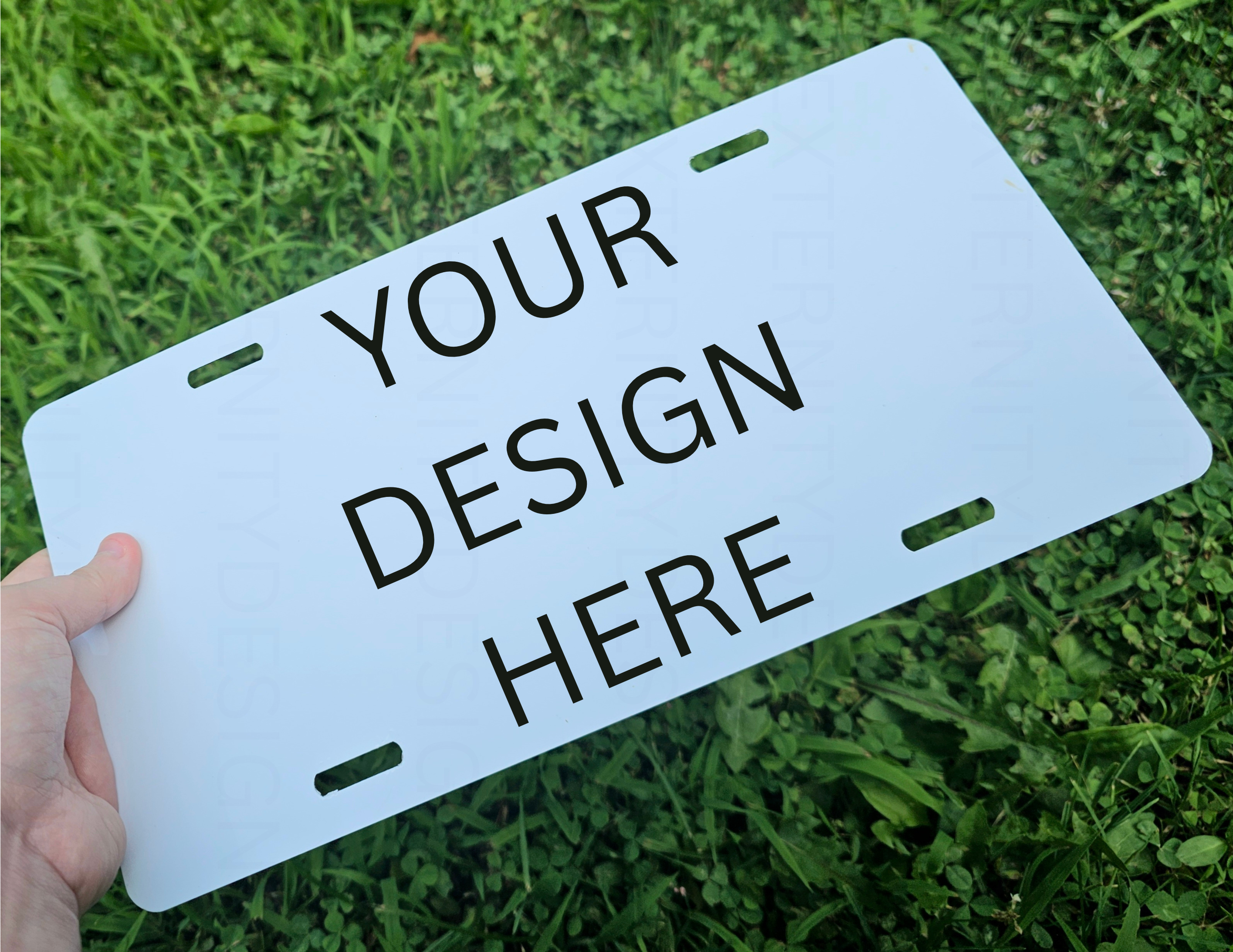 White license plate with 'Your Design Here' text held over grass