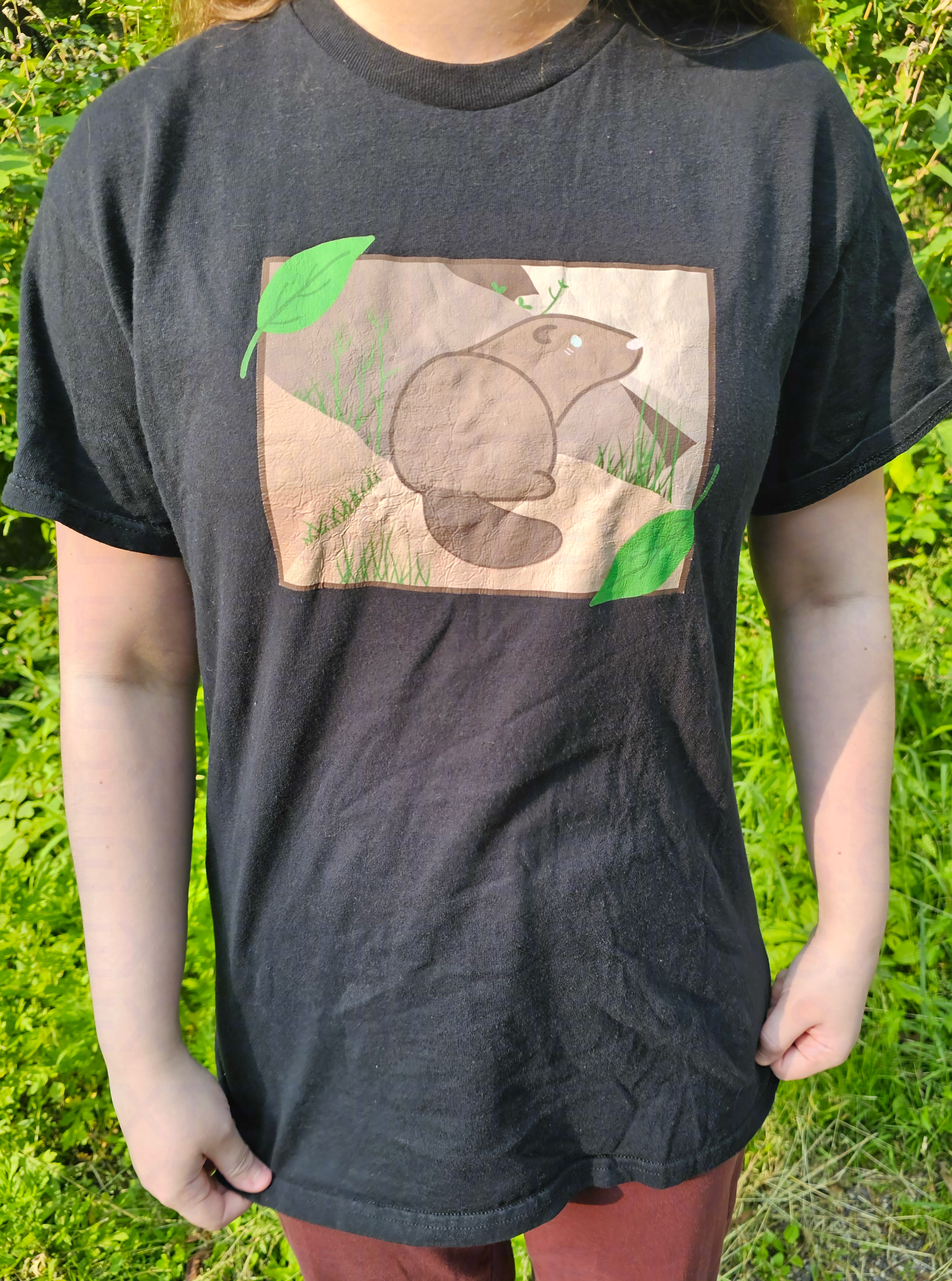 A person wearing a black t-shirt with a graphic design of a groundhog and leaves outdoors.