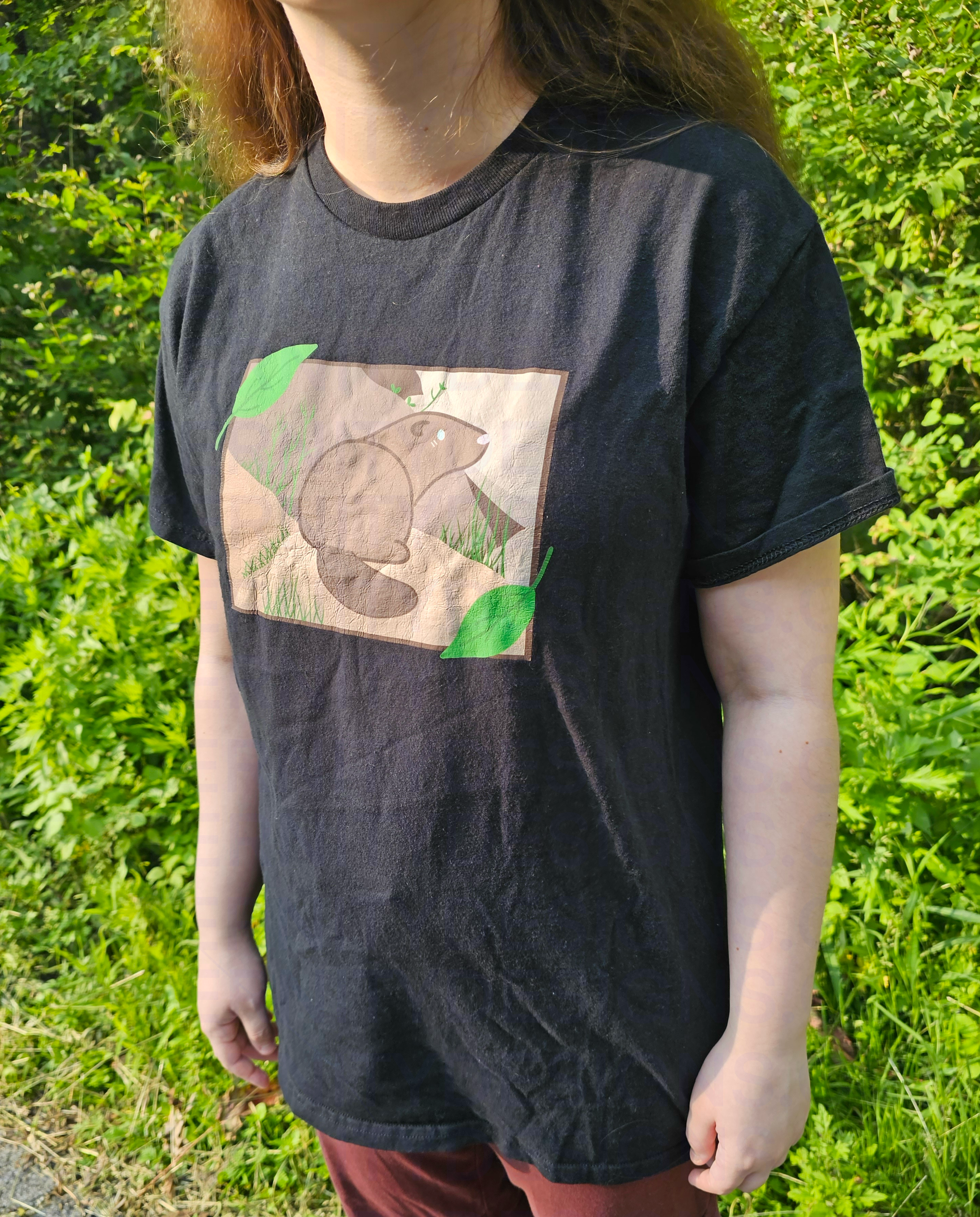 A person wearing a black t-shirt with a graphic design of a groundhog and leaves outdoors.