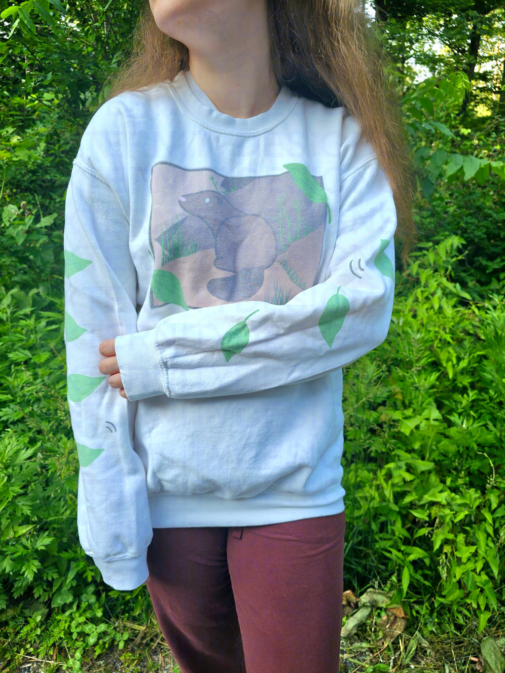 A person with their left arm across their stomach wearing a white sweatshirt featuring a groundhog drawing on the chest and leaves on the sleeves.