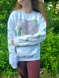 A person with their left arm across their stomach wearing a white sweatshirt featuring a groundhog drawing on the chest and leaves on the sleeves.