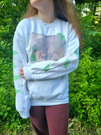 A person with their left arm across their stomach wearing a white sweatshirt featuring a groundhog drawing on the chest and leaves on the sleeves.