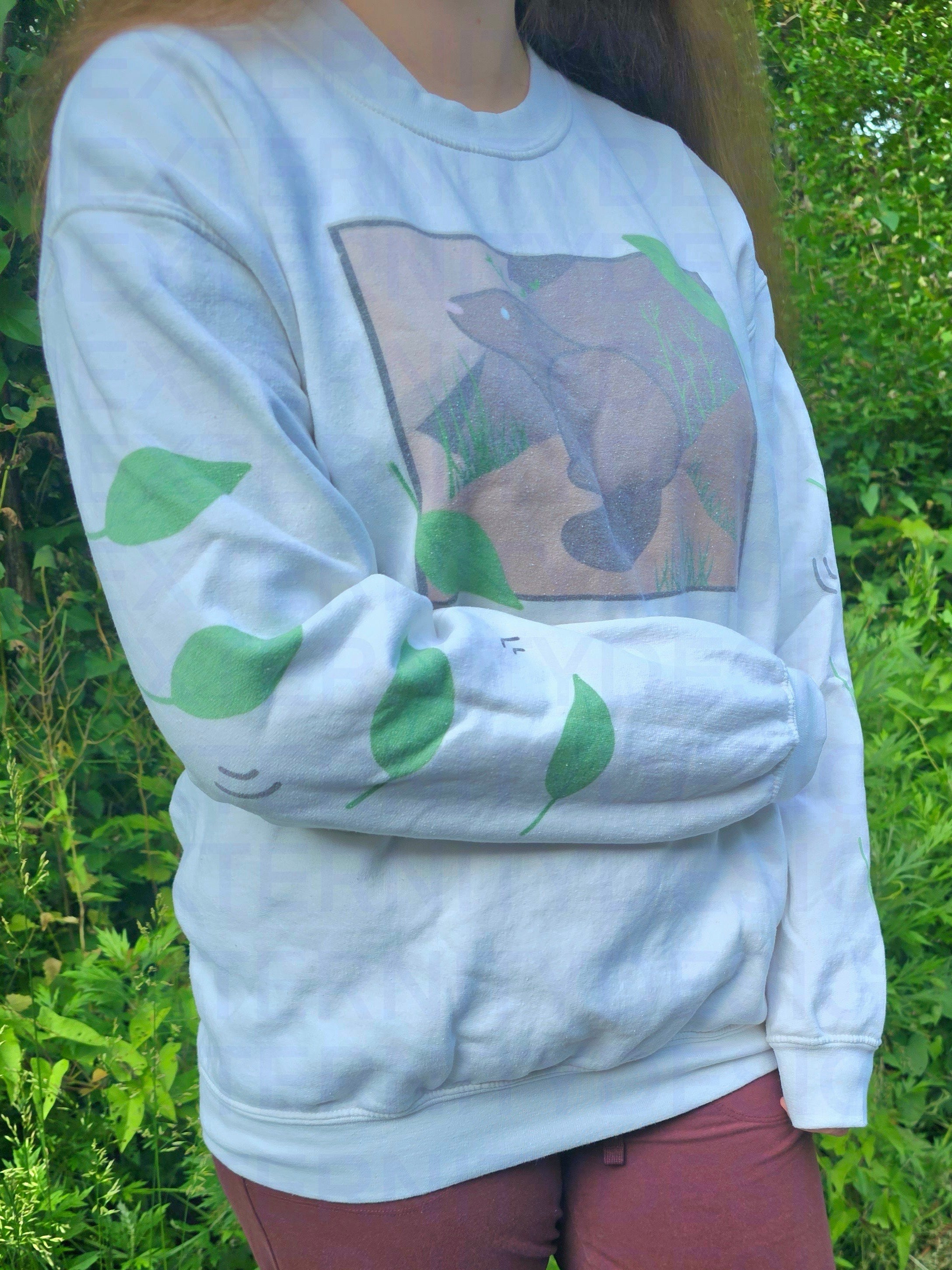 A person with their right arm across their stomach wearing a white sweatshirt featuring a groundhog drawing on the chest and leaves on the sleeves.