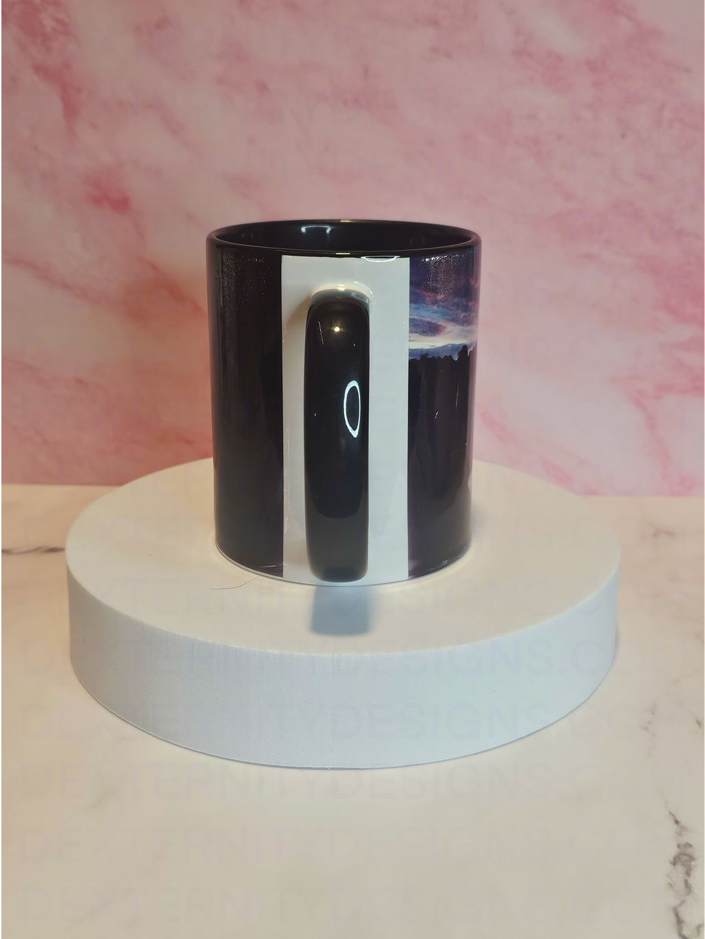 Back view of a mug with a scenic sunrise design on a white platform against a pink marble background