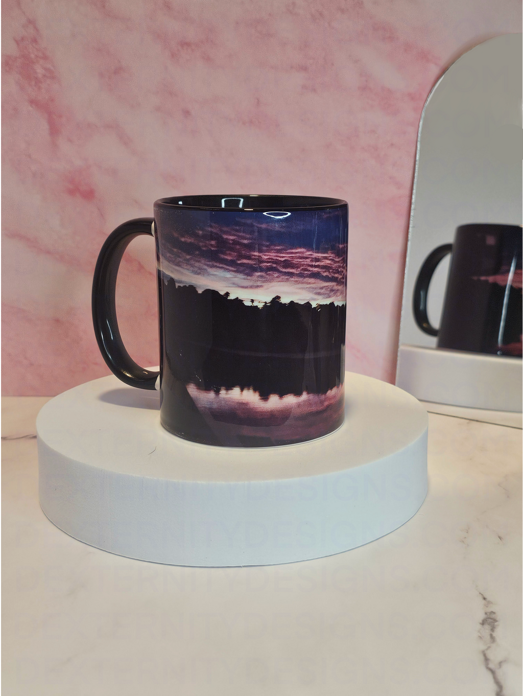 Mug with a scenic sunrise design on a white platform against a pink marble background with a mirror reflecting it's back behind it.