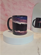 Mug with a scenic sunrise design on a white platform against a pink marble background with a mirror reflecting it's back behind it.