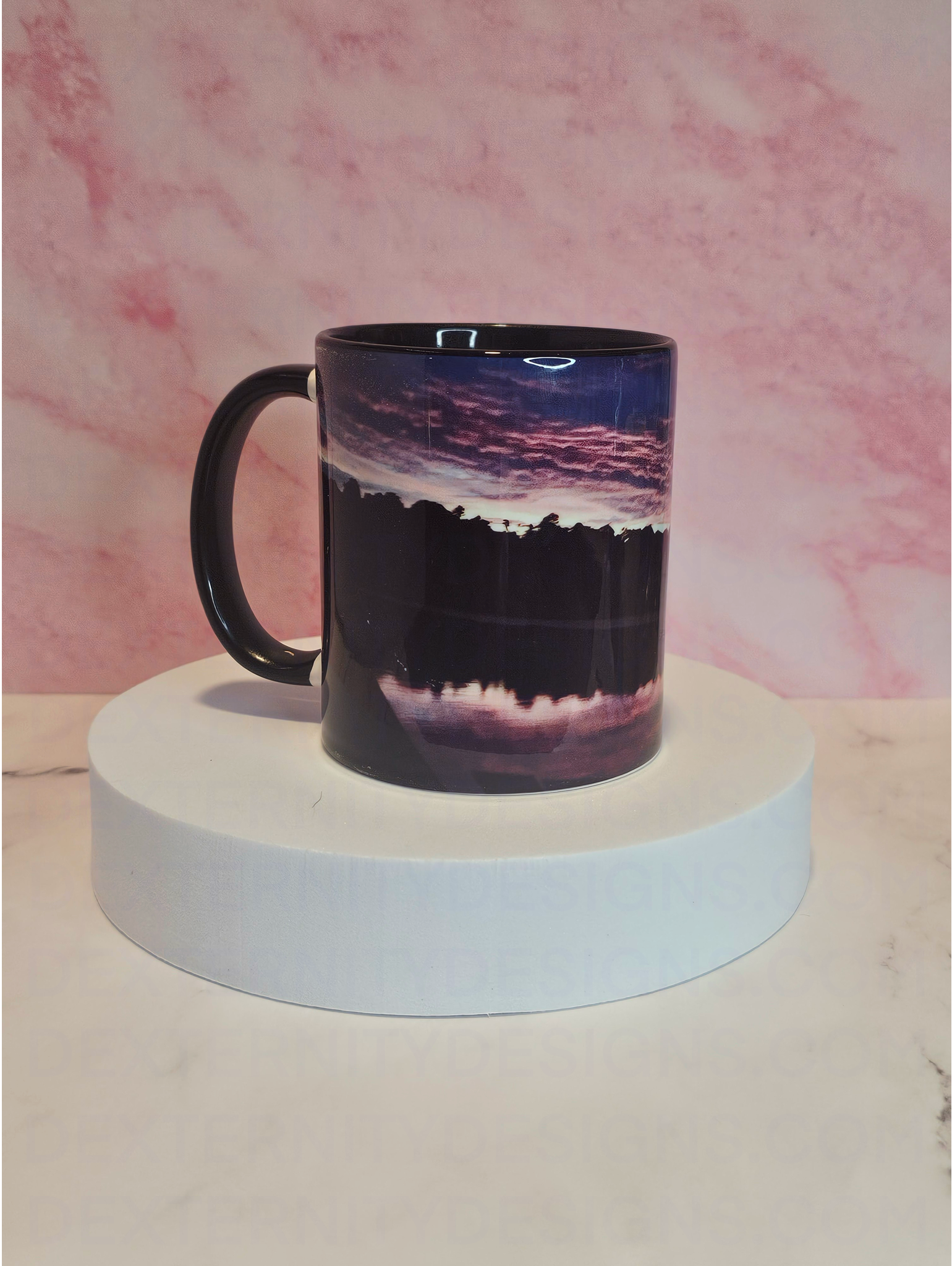 Side view of a mug with a scenic sunrise design on a white platform against a pink marble background