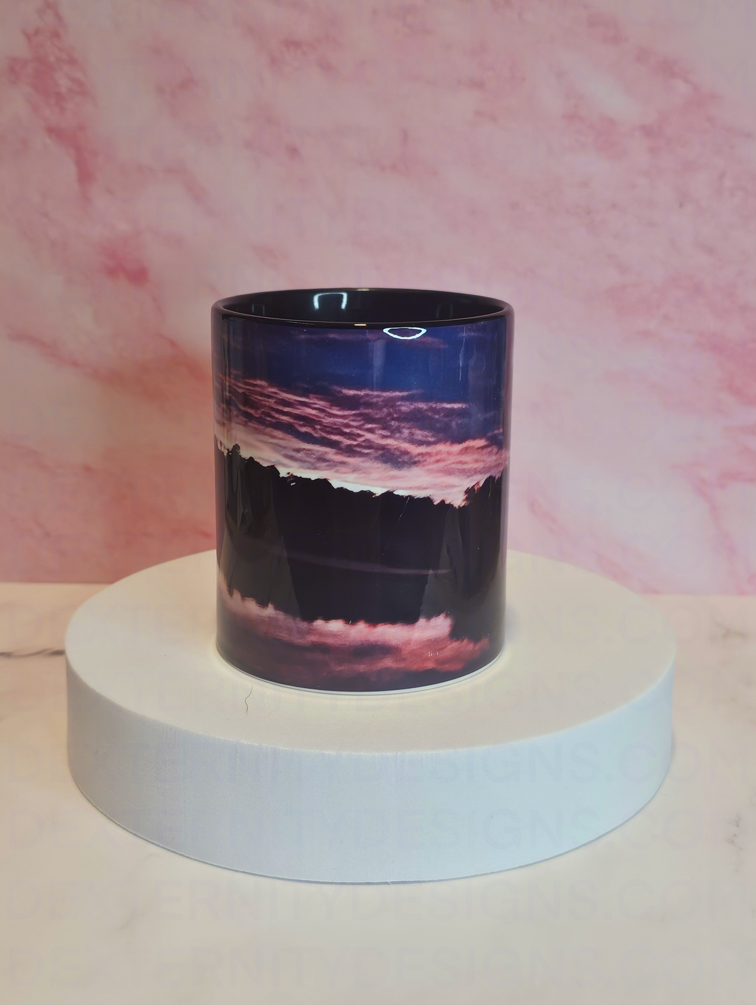 Front view of a mug with a scenic sunrise design on a white platform against a pink marble background